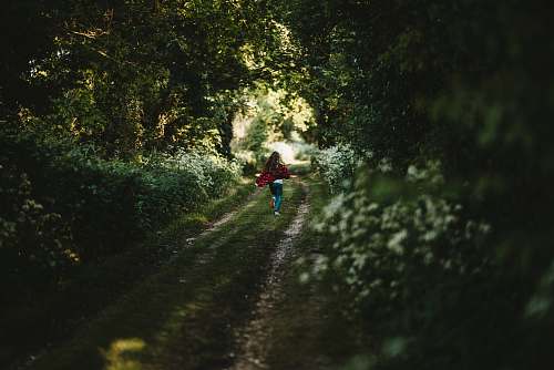 sports woman in red shirt and green pants walking on grass road between trees during daytime tree